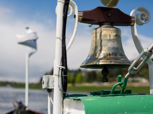 Service & Info Auf einem Schiff hängt die goldene Schiffsglocke. Im Hintergrund sind verschwommen der Fluss und grüne Bäume zu sehen.